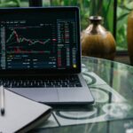 Laptop displaying cryptocurrency stocks and graphs on a glass table with a notepad.